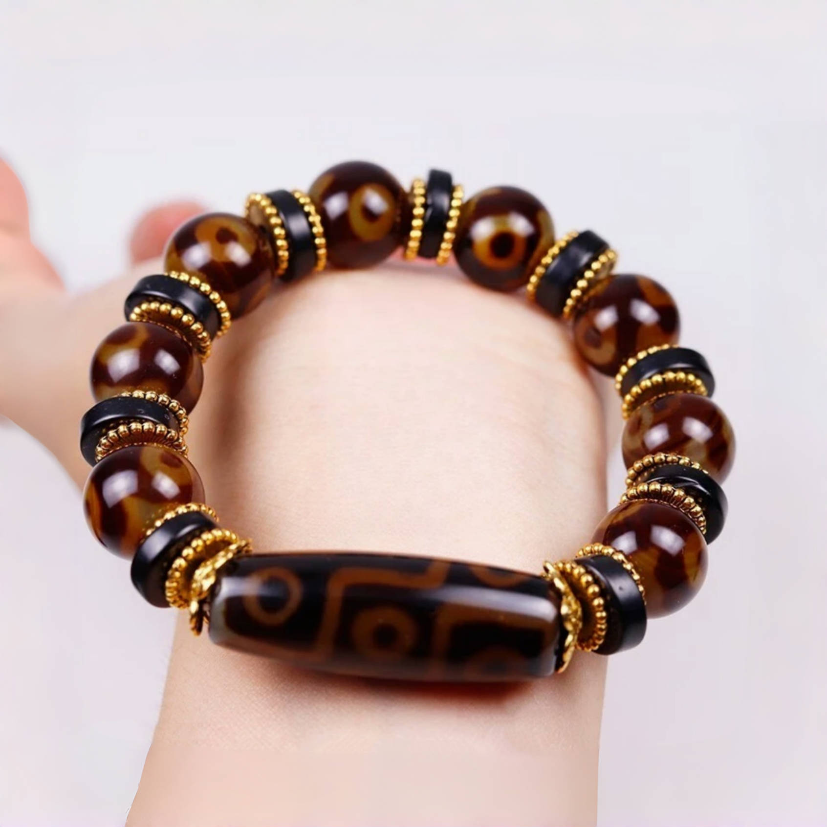 The bracelet is worn around a wrist, offering a clear view of how it looks when worn. The central Dzi bead sits prominently at the top, while the polished brown agate beads and gold-accented black spacers encircle the wrist. The image highlights the bracelet’s flexibility, rich tones, and bold spiritual design.