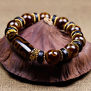 The bracelet is gracefully arranged on a stylized, mountain-shaped wooden prop. The elongated Dzi bead with its nine-eye motif rests prominently at the front, flanked by brown agate beads and gold-rimmed black spacers. The earthy tones and organic backdrop evoke a sense of spiritual grounding and heritage artistry.