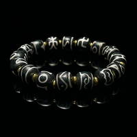 A high-contrast luxury presentation of a Tibetan-style Dzi bead bracelet laid on a glossy black mirror background. The reflection enhances the deep black tones of the beads, which are etched with intricate white script. Gold-tone spacers add a touch of elegance, making this a visually striking and spiritually meaningful accessory.