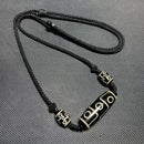 The necklace showcases a Tibetan Nine-Eyed Dzi bead surrounded by smaller beads, strung on a knotted black cord, emphasizing its traditional design.