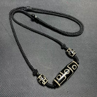 The necklace showcases a Tibetan Nine-Eyed Dzi bead surrounded by smaller beads, strung on a knotted black cord, emphasizing its traditional design.