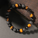 Bracelet resting on textured wood, composed of tiger’s eye, black obsidian, and porous lava beads, anchored by a silver cross charm.