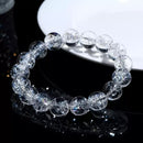 A clear quartz bracelet laid flat beside a white dish on black surface, capturing rainbow refractions within the beads.