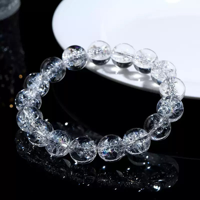 A clear quartz bracelet laid flat beside a white dish on black surface, capturing rainbow refractions within the beads.