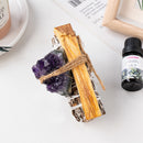 A white sage and palo santo bundle tied with raw amethyst crystal, placed beside essential oil and candle.