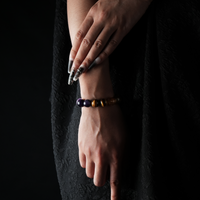 A stylish model with manicured nails wearing a gemstone bracelet featuring purple amethyst and golden tiger eye beads, contrasted against a black textured outfit and dark backdrop