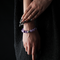 A close-up of a woman's hands gracefully posed while wearing a round-beaded amethyst bracelet. The bracelet features polished purple stones and a central silver-toned bead with intricate detailing. The model is dressed in a textured black outfit, with dramatic black-and-silver nail art, set against a dark background.