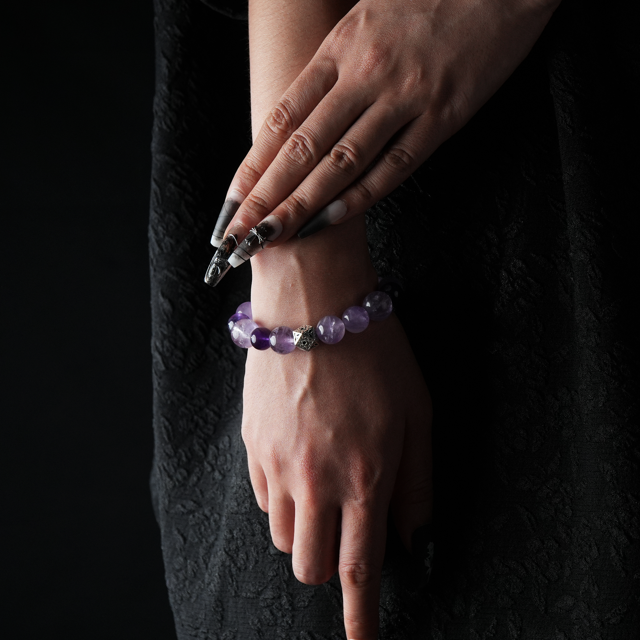 A close-up of a woman's hands gracefully posed while wearing a round-beaded amethyst bracelet. The bracelet features polished purple stones and a central silver-toned bead with intricate detailing. The model is dressed in a textured black outfit, with dramatic black-and-silver nail art, set against a dark background.