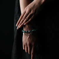 A wrist adorned with a dark green cracked energy bead bracelet featuring a silver feather charm and dainty chain, embodying forest strength and intuitive calm.
