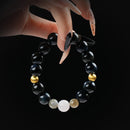 Black obsidian bracelet with gold hematite and white moonstone beads, held by hand on black background for a powerful and protective look.