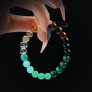 Bracelet composed of various gemstone beads in green, black, yellow, and brown shades, held by hand against a black background.