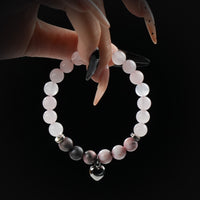 Delicate bracelet made of rose quartz and pink-toned beads, featuring a metallic heart charm, held by hand against a black background.