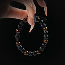 Tiger's eye, lava stone, and obsidian beaded bracelet with black cross charm, held by hand on black background for strength and faith.